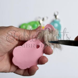 May include: A pink leather keychain with a scalloped edge is being assembled. A pair of tweezers is used to trim the light brown fur attached to the keychain. Other keychains in green and blue are in the background.