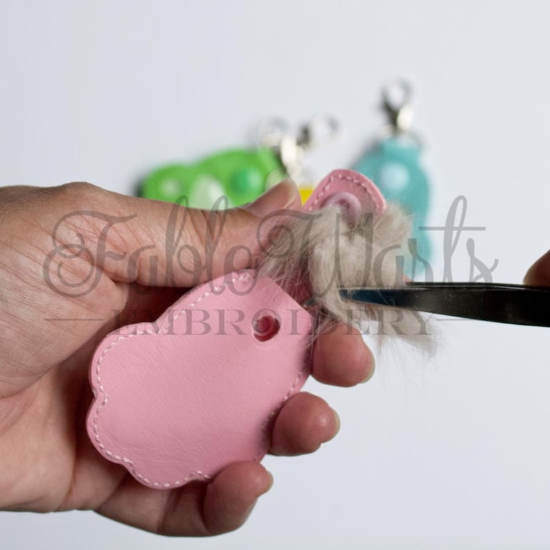 May include: A pink leather keychain with a scalloped edge is being assembled. A pair of tweezers is used to trim the light brown fur attached to the keychain. Other keychains in green and blue are in the background.