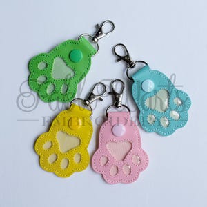 May include: Four colorful paw-shaped keychains in green, yellow, blue, and pink. Each keychain has a silver clasp and a button detail. The paw pads are visible through a clear, heart-shaped window.