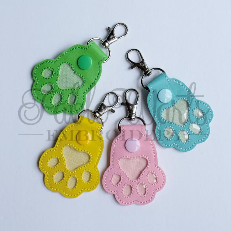 May include: Four colorful paw-shaped keychains in green, yellow, blue, and pink. Each keychain has a silver clasp and a button detail. The paw pads are visible through a clear, heart-shaped window.