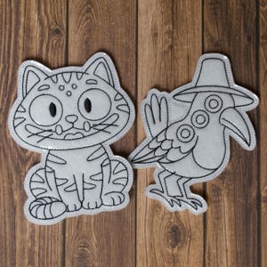 May include: Two white fabric patches with black stitched outlines. One is a cartoon cat with large eyes and stripes. The other is a bird wearing a hat with a long beak. Both are on a wooden surface.