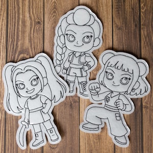 Puede incluir: Tres parches de tela blanca con contornos bordados en negro de personajes de dibujos animados. Cada parche representa una figura femenina diferente en una pose dinámica, con rasgos faciales y ropa detallados. Los parches están dispuestos sobre una superficie de madera.