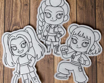 Puede incluir: Tres parches de tela blanca con contornos bordados en negro de personajes de dibujos animados. Cada parche representa una figura femenina diferente en una pose dinámica, con rasgos faciales y ropa detallados. Los parches están dispuestos sobre una superficie de madera.