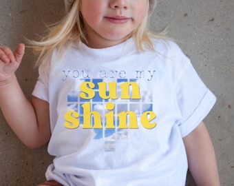 Może przedstawiać: Biała koszulka z niebiesko-żółtym nadrukiem z napisem "you are my sunshine".