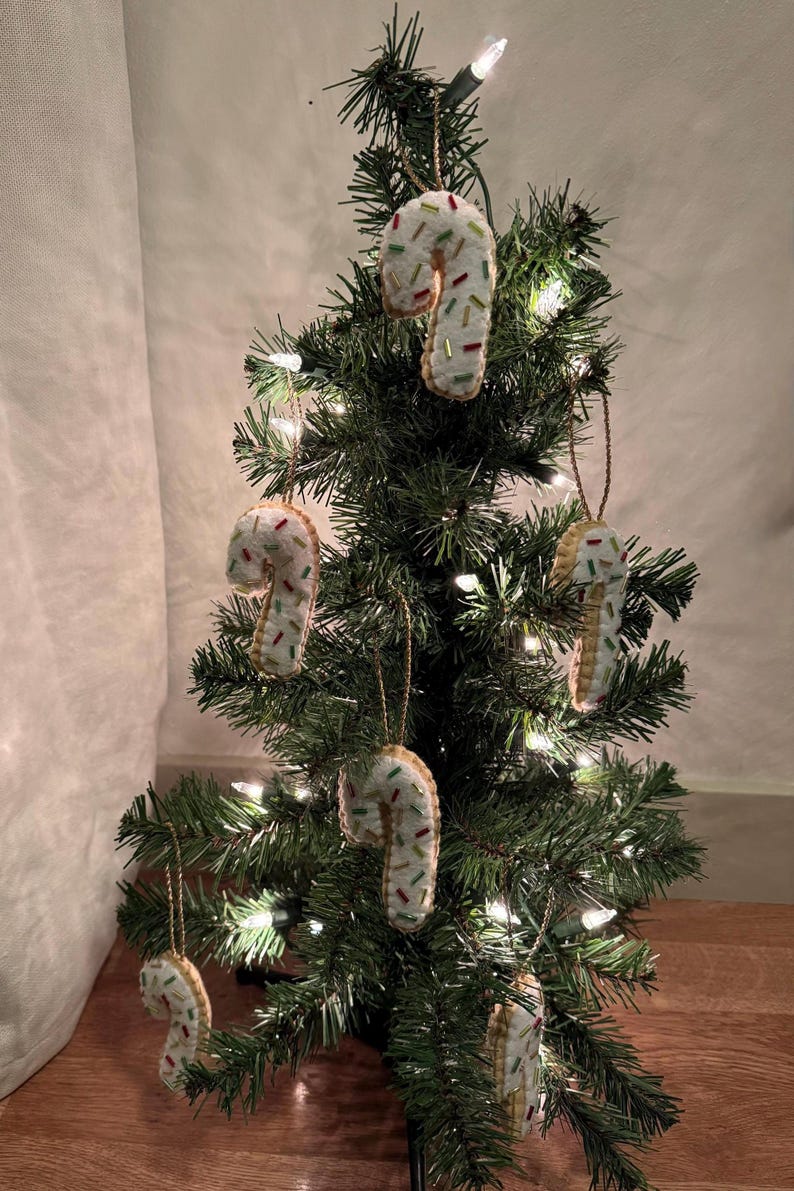 May include: A small Christmas tree decorated with handmade candy cane ornaments. The candy canes are white with colorful sprinkles and hang from the tree with gold string. The tree is lit with small white lights.