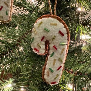 May include: A felt candy cane Christmas ornament with a white base and brown trim. The ornament is decorated with red, green, and gold beads, and hangs from a gold string. The ornament is hanging on a Christmas tree.