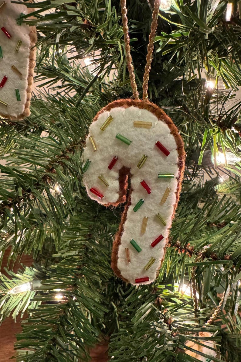 May include: A felt candy cane Christmas ornament with a white base and brown trim. The ornament is decorated with red, green, and gold beads, and hangs from a gold string. The ornament is hanging on a Christmas tree.