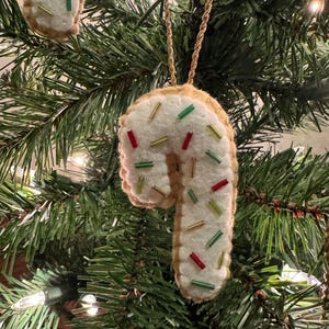 May include: A felt candy cane Christmas ornament with white icing and colorful sprinkles. The ornament is trimmed with a gold cord for hanging and is displayed on a Christmas tree with green needles and white lights.