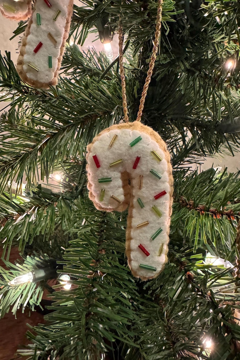May include: A felt candy cane Christmas ornament with white icing and colorful sprinkles. The ornament is trimmed with a gold cord for hanging and is displayed on a Christmas tree with green needles and white lights.