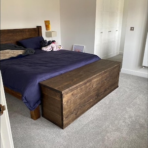 K&ouml;nnte beinhalten: Ein dunkles braunes Holzbettgestell mit einem blauen Bettbezug und einem passenden dunklen braunen Holzfu&szlig;teil. Das Bett steht in einem Schlafzimmer mit wei&szlig;en W&auml;nden und einem wei&szlig;en Kleiderschrank.