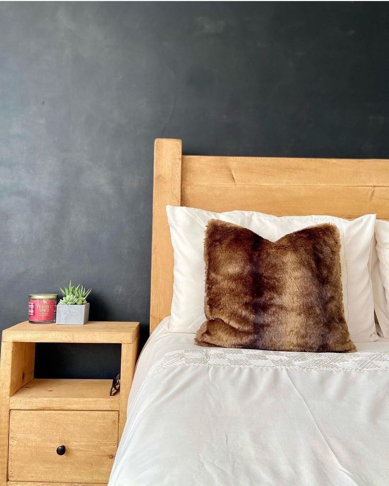 May include: A wooden bed frame with a white duvet and a brown faux fur pillow. A small wooden nightstand with a candle and two succulents sits next to the bed.