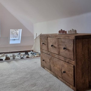 May include: A rustic wooden dresser with six drawers and black metal handles. The dresser is made of reclaimed wood and has a natural finish. It is sitting in a room with a window and a carpeted floor.