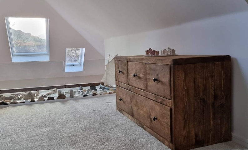 May include: A rustic wooden dresser with six drawers and black metal handles. The dresser is made of reclaimed wood and has a natural finish. It is sitting in a room with a window and a carpeted floor.