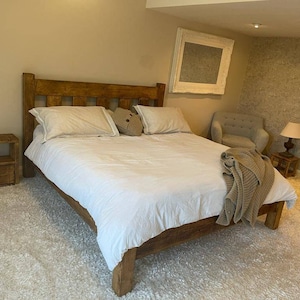 May include: A rustic wooden bed frame with a white duvet and pillows. The bed is in a bedroom with a white rug, a wooden nightstand, and a chair.