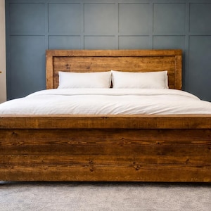 Puede incluir: Una cama de madera r&uacute;stica con cabecero y piecero. La cama est&aacute; hecha de madera te&ntilde;ida oscura y tiene un dise&ntilde;o rectangular simple. La cama est&aacute; cubierta con ropa de cama y almohadas blancas. El fondo es una pared azul oscuro.