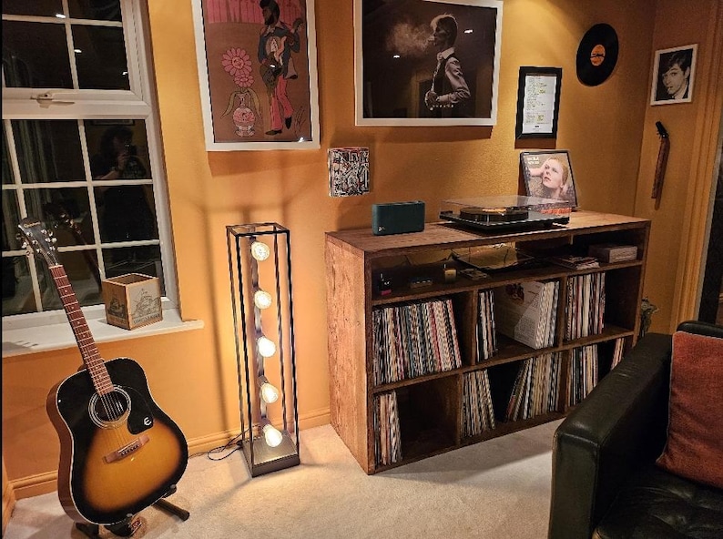 May include: A wooden record shelf with a turntable on top. The shelf has six shelves and is filled with vinyl records. There is a guitar leaning against the shelf and a floor lamp with five light bulbs.