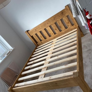 May include: A wooden bed frame with a slatted headboard and footboard. The frame is made of natural wood with a light brown finish. The slats are made of a lighter wood and are visible through the gaps in the headboard.