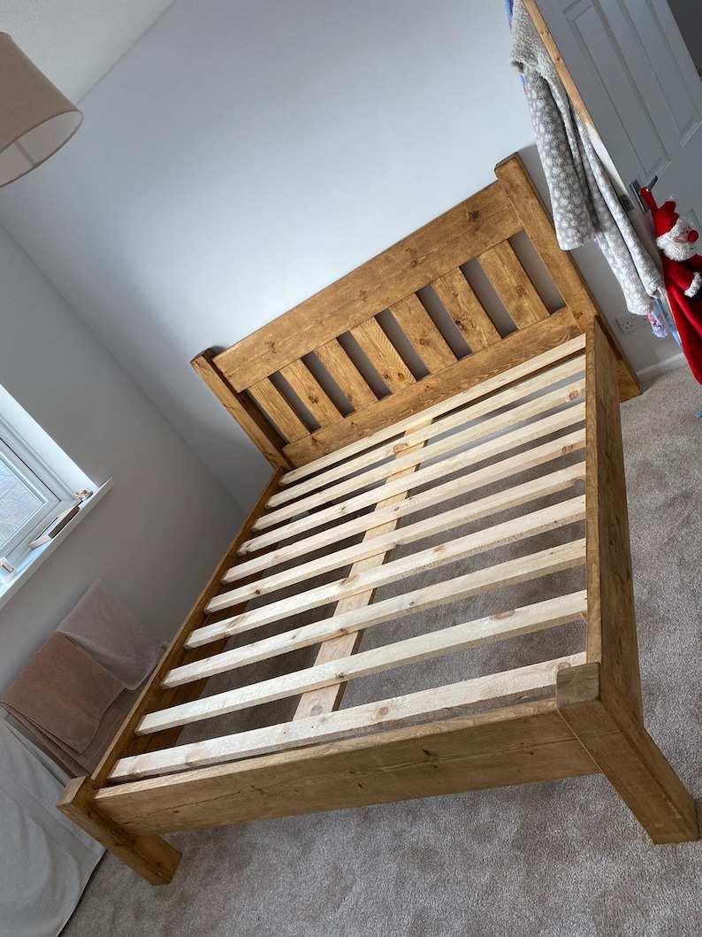 May include: A wooden bed frame with a slatted headboard and footboard. The frame is made of natural wood with a light brown finish. The slats are made of a lighter wood and are visible through the gaps in the headboard.