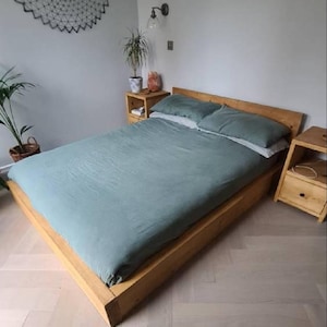 May include: A wooden platform bed frame with a light green duvet and two pillows. The bed is in a bedroom with light wood floors and a white wall.