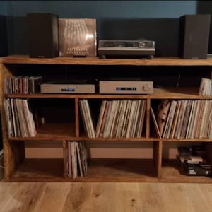May include: A wooden record player stand with six shelves, filled with vinyl records. Two speakers and a record player are on top of the stand.