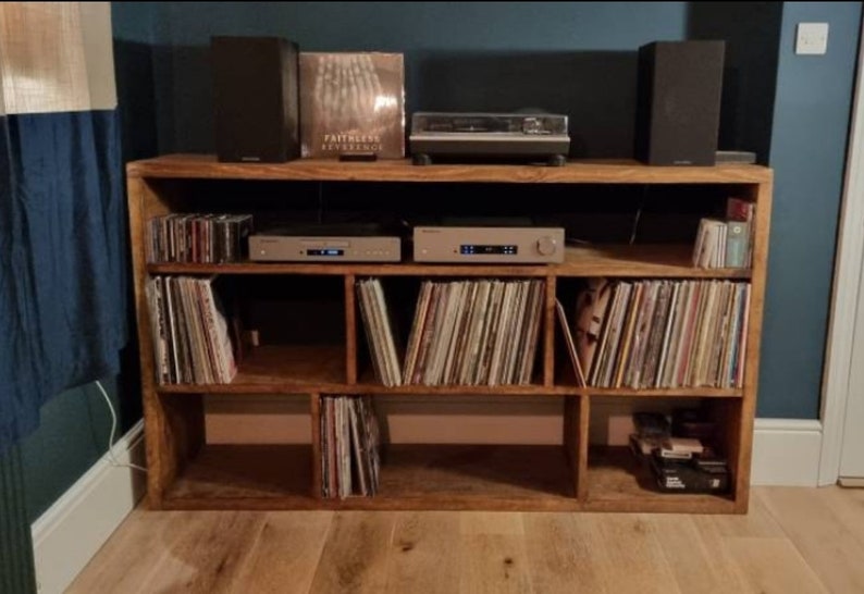 May include: A wooden record player stand with six shelves, filled with vinyl records. Two speakers and a record player are on top of the stand.
