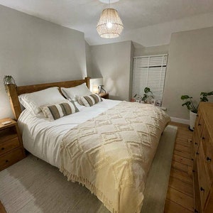 Puede incluir: Un dormitorio con una estructura de cama de madera y un edred&oacute;n blanco. La cama tiene una manta de tiro beige con un patr&oacute;n de diamante. Hay dos mesitas de noche a cada lado de la cama, cada una con una l&aacute;mpara. La habitaci&oacute;n tiene una pared gris claro y un suelo de madera.