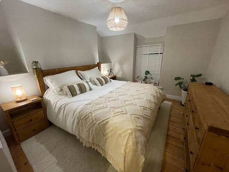Puede incluir: Un dormitorio con una estructura de cama de madera y un edred&oacute;n blanco. La cama tiene una manta de tiro beige con un patr&oacute;n de diamante. Hay dos mesitas de noche a cada lado de la cama, cada una con una l&aacute;mpara. La habitaci&oacute;n tiene una pared gris claro y un suelo de madera.