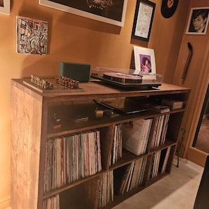 May include: A wooden record player stand with multiple shelves filled with vinyl records. The stand has a light brown wood finish and is in a room with a light brown wall.