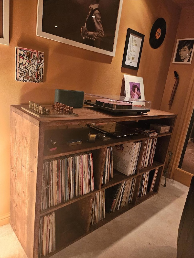 May include: A wooden record player stand with multiple shelves filled with vinyl records. The stand has a light brown wood finish and is in a room with a light brown wall.