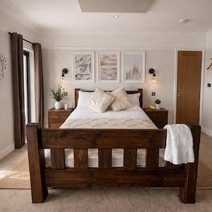 May include: A dark wooden bed frame is the focal point of the bedroom. The bed is made with white bedding and fluffy pillows. Two matching bedside tables with lamps and decor sit either side of the bed. Three framed landscape prints hang above the bed.