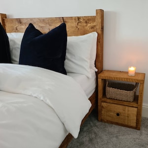 May include: A wooden bed frame with a white duvet and black pillows. A wooden nightstand with a drawer and a wicker basket sits next to the bed.