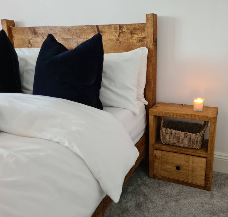 May include: A wooden bed frame with a white duvet and black pillows. A wooden nightstand with a drawer and a wicker basket sits next to the bed.