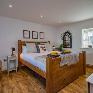 May include: A bedroom with a wooden bed frame, white bedding, and decorative pillows. A wicker chair, side table with a lamp, and framed artwork are also visible. The room has hardwood floors and a window with blinds.