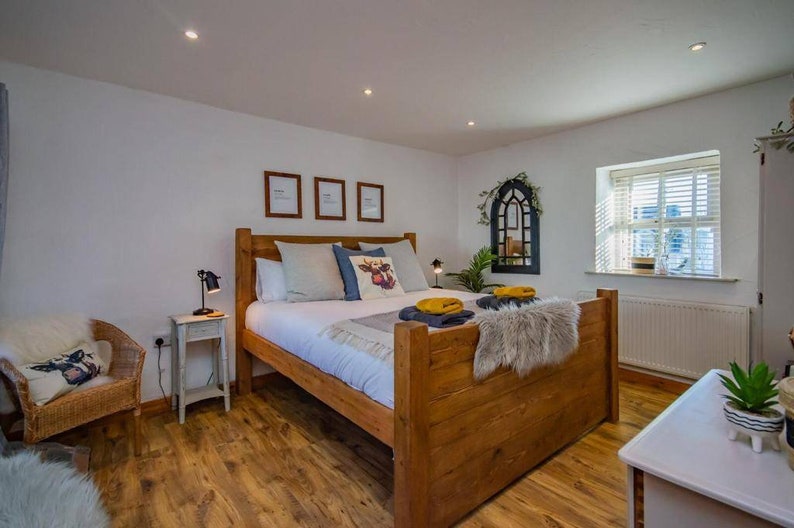 May include: A bedroom with a wooden bed frame, white bedding, and decorative pillows. A wicker chair, side table with a lamp, and framed artwork are also visible. The room has hardwood floors and a window with blinds.