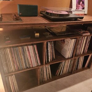 May include: A wooden record player stand with multiple shelves filled with vinyl records. The stand has a turntable on top and a small speaker on the left side.