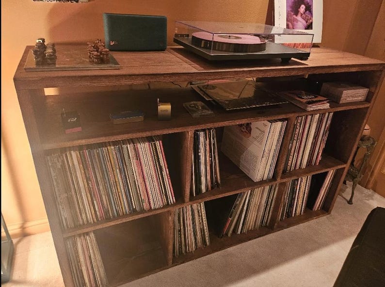 May include: A wooden record player stand with multiple shelves filled with vinyl records. The stand has a turntable on top and a small speaker on the left side.