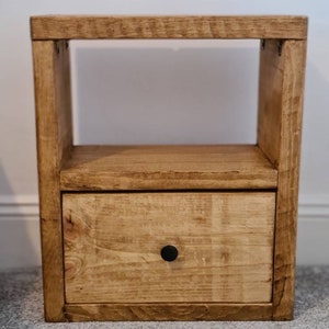 May include: A wooden bedside table with a single drawer and a shelf. The table is stained a light brown color and has a black knob on the drawer.
