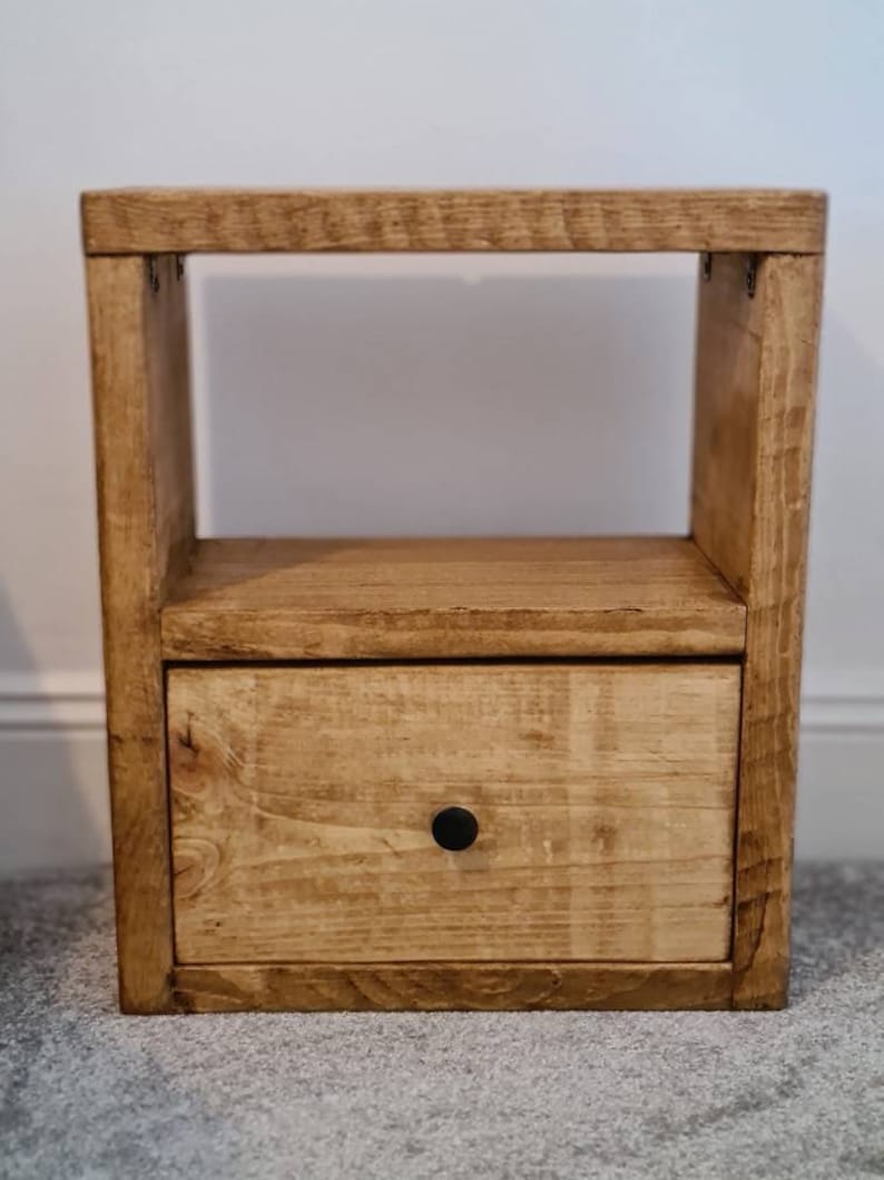 May include: A wooden bedside table with a single drawer and a shelf. The table is stained a light brown color and has a black knob on the drawer.