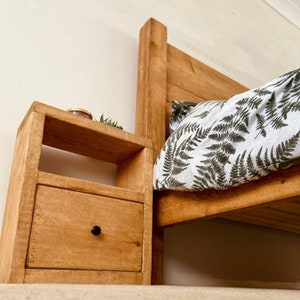 May include: A wooden bedside table with a drawer and a shelf. The table is stained a light brown color and has a black knob on the drawer. The table is next to a bed with a white and green patterned duvet cover.