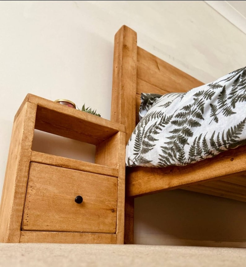 May include: A wooden bedside table with a drawer and a shelf. The table is stained a light brown color and has a black knob on the drawer. The table is next to a bed with a white and green patterned duvet cover.