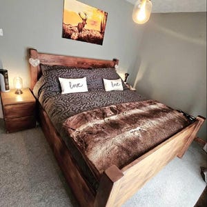 May include: A wooden bed frame with a brown patterned comforter and a faux fur throw. Two white pillows with the word "love" are on the bed. A landscape painting hangs above the bed, and a bedside table with a lamp is on the left.