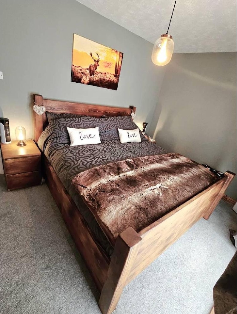 May include: A wooden bed frame with a brown patterned comforter and a faux fur throw. Two white pillows with the word "love" are on the bed. A landscape painting hangs above the bed, and a bedside table with a lamp is on the left.