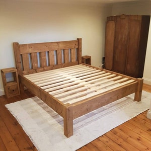 Puede incluir: Un marco de cama de madera r&uacute;stica con una cabecera y un piecero de listones. El marco de la cama est&aacute; hecho de madera recuperada y tiene un acabado natural. El marco de la cama est&aacute; sobre una alfombra blanca en una habitaci&oacute;n con suelo de madera.