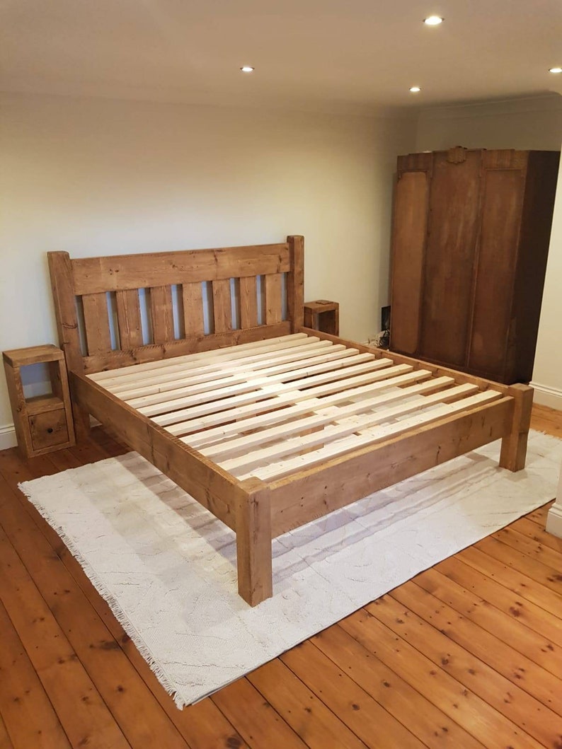 Puede incluir: Un marco de cama de madera r&uacute;stica con una cabecera y un piecero de listones. El marco de la cama est&aacute; hecho de madera recuperada y tiene un acabado natural. El marco de la cama est&aacute; sobre una alfombra blanca en una habitaci&oacute;n con suelo de madera.