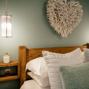 May include: A bedroom scene featuring a wooden headboard and a floating nightstand. White pillows and a textured green pillow rest on the bed. A decorative heart made of white branches hangs on the wall, and a pendant light hangs from the ceiling.