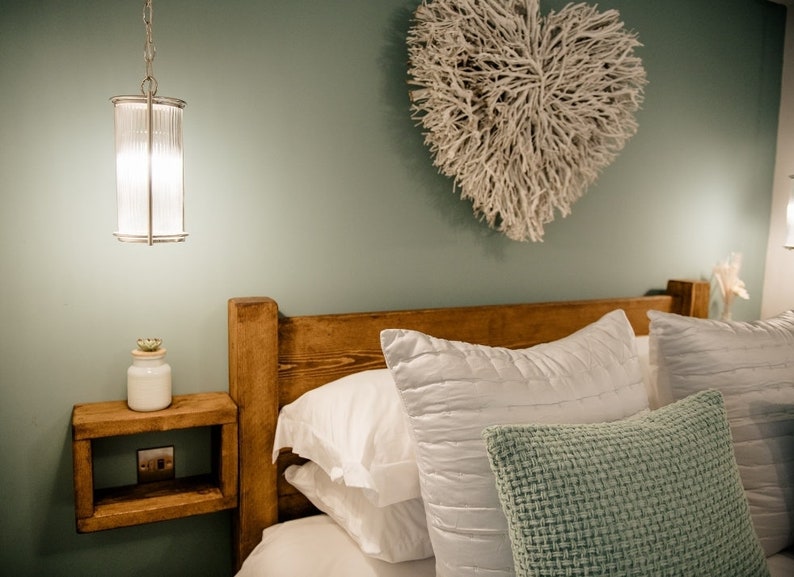 May include: A bedroom scene featuring a wooden headboard and a floating nightstand. White pillows and a textured green pillow rest on the bed. A decorative heart made of white branches hangs on the wall, and a pendant light hangs from the ceiling.