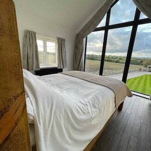 May include: A white bed with a wooden headboard and a beige throw blanket. The bed is in a bedroom with a large window overlooking a green field.