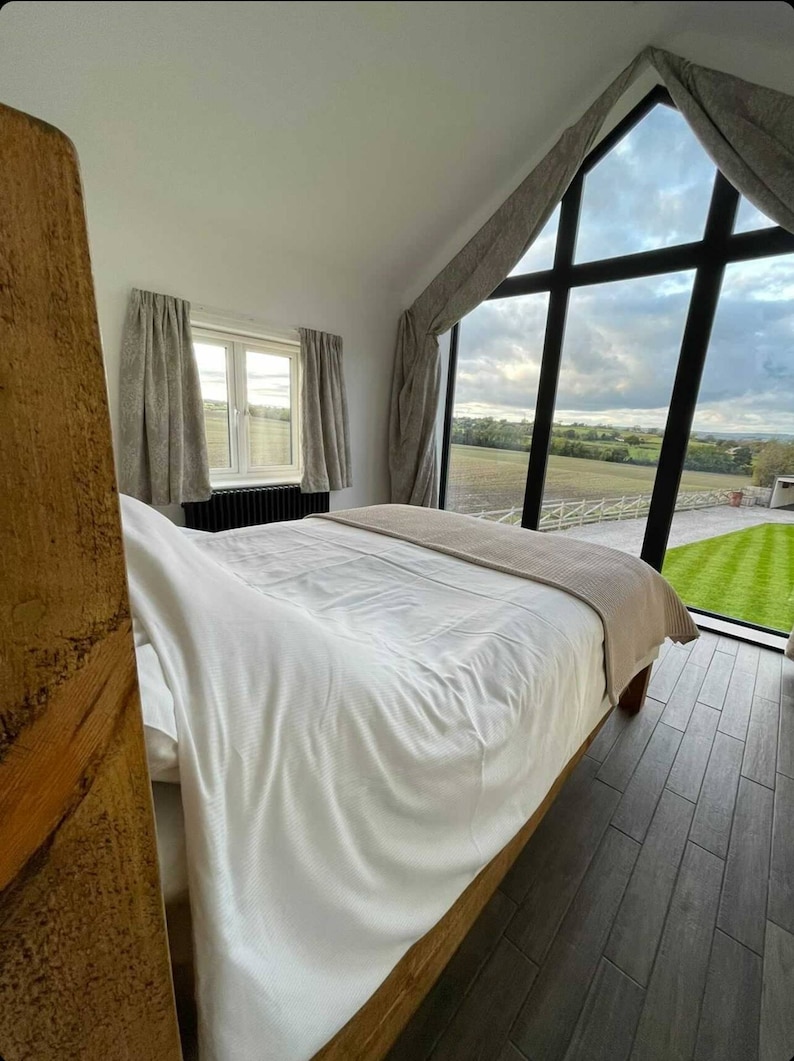 May include: A white bed with a wooden headboard and a beige throw blanket. The bed is in a bedroom with a large window overlooking a green field.