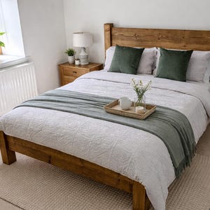 May include: A wooden bed with a headboard, dressed with white bedding, green cushions, and a grey throw. A tray with a mug, flowers, and candles sits on the bed. A rug and a bedside table with a lamp are also visible.