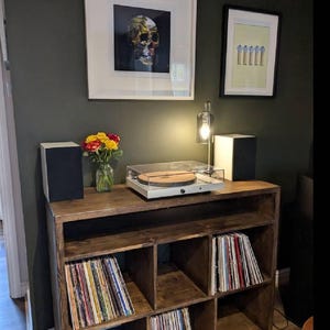 May include: A dark wood record player stand with six shelves, filled with vinyl records. A turntable sits on the top shelf, with a framed print of a skull above it.